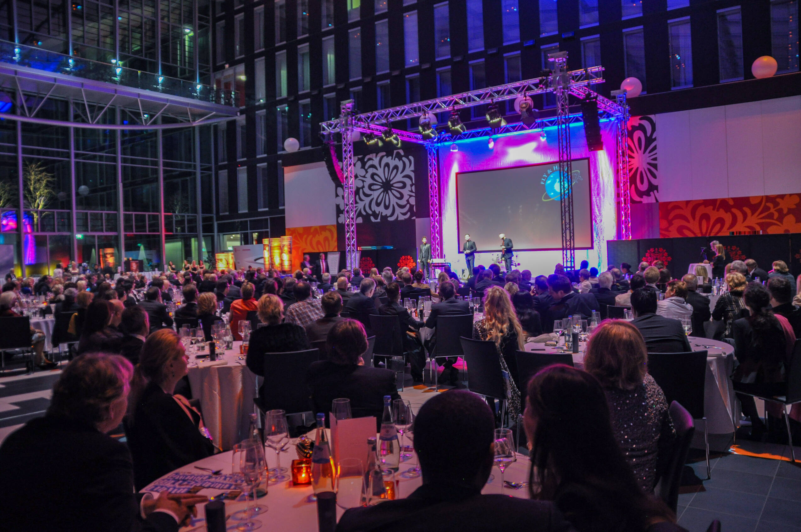 Menschen bei Gala Veranstaltung, FLY&HELP Gala, Veranstaltung mit beleuchteter Bühne und Menschen an Tischen, Feierlichkeit mit vielen Menschen in Abendkleidung